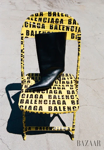 High-heeled leather boot is propped on a yellow folding chair with black BALENCIAGA logos in a fashion advertisement.