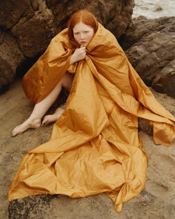 Red-haired woman wrapped tightly in an ochre tarp sitting on a sandy beach against rocks