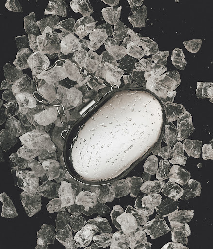 Close-up of a metallic wristlet purse with a chain handle, covered in water droplets, resting on a bed of small, clear ice chunks.