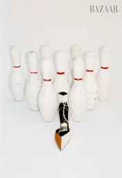 A stiletto shoe positioned in front of a triangular arrangement of white bowling pins against a seamless white background.