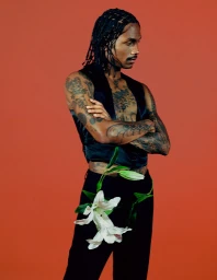 Man with braided hair and tattoos wearing a black vest and trousers with white lilies against an orange backdrop