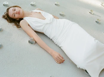 A model in a white gown lies on a light gray textured surface surrounded by small clear glass decorative objects.