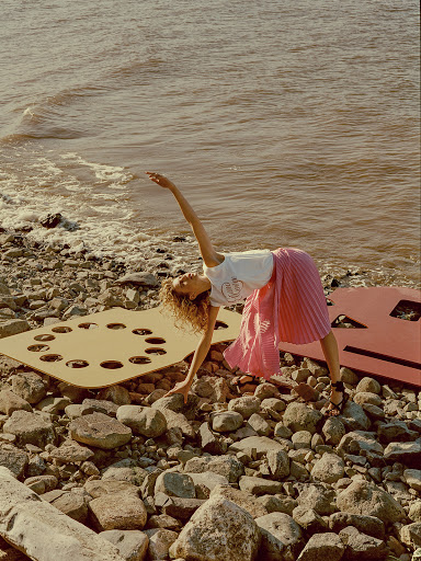 Woman performs yoga on a rocky beach in a pink skirt with the ocean in the background. The photograph is in landscape orientation.