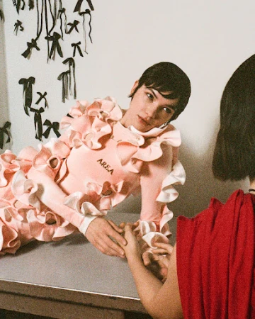 Two fair-skinned people in a photo studio. One is wearing a pink dress with ruffles, and the other is wearing a red dress.