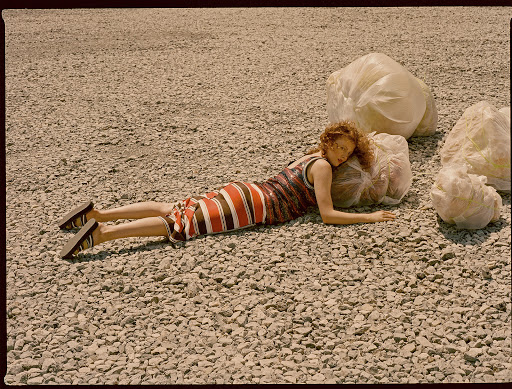 A woman with curly red hair poses on a gravel surface next to large, white plastic bags. She is wearing a brown and orange striped dress.