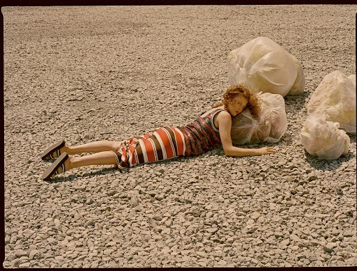 A woman with curly red hair poses on a gravel surface next to large, white plastic bags. She is wearing a brown and orange striped dress.