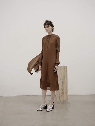 Fashion shot of a woman in a long-sleeved brown dress and white heeled shoes standing in front of a neutral wall and wooden stand.