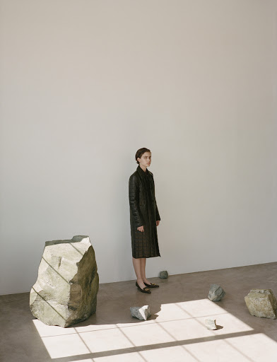 Woman in dark quilted coat standing in a minimalist room with sculptural rocks and window light patterns on the floor.