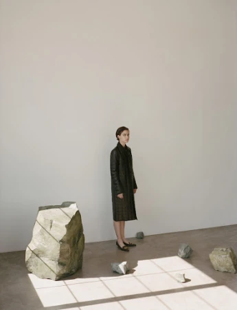 Woman in dark quilted coat standing in a minimalist room with sculptural rocks and window light patterns on the floor.