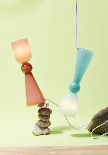Two colorful glass lamps, one peach and one turquoise, are propped up by rocks, with a light green background and a wooden surface.
