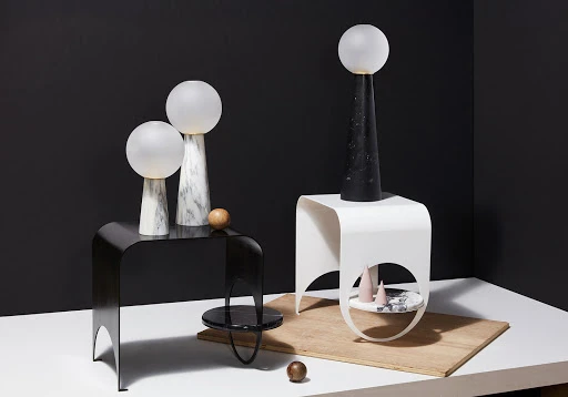 Contemporary interior showcasing two table lamps with spherical shades on marble bases, set on modern black and white tables against a dark background.