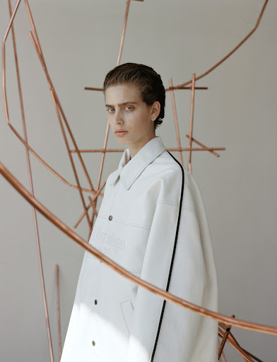 An androgynous-looking person with copper pipe art poses in a white jacket with black trim, medium studio shot with soft diffused light.