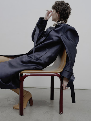 Woman in draped navy coat and heeled boots sits on a chair in a minimalist photo studio against a white backdrop.