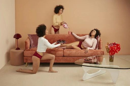 Three women in an indoor setting with peach-toned colors. One kneels holding a tray of cupcakes, another pours from a tea pot, the third reclines.