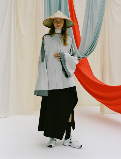A model poses indoors wearing a pale blue top, black skirt, New Balance sneakers, and a Asian-style conical hat in a fashion editorial.