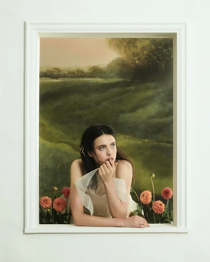 Woman in a white dress and tulle shawl gazes upward near dahlia flowers, framed by a white window against a painted landscape backdrop.