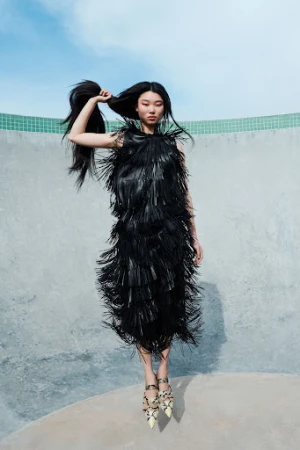 Model in black fringe dress holding long dark hair against a concrete pool backdrop with clear blue sky