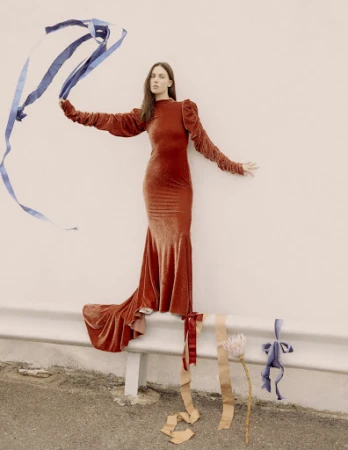 Model in a long rust-orange velvet dress holding blue ribbons against an outdoor concrete barrier by set designer Lauren Bahr.