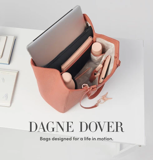 Salmon-colored Dagne Dover tote bag on a white surface, open to reveal a laptop, journal, cosmetics, and other personal items inside.