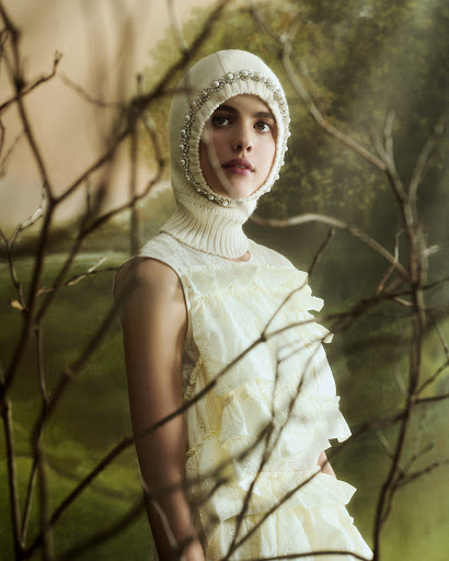 Fashion portrait of a woman wearing a knitted balaclava with pearl trim and a ruffled lace dress, posing against a painterly background.