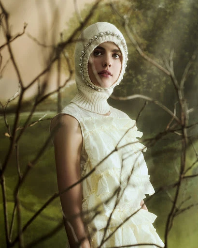 Fashion portrait of a woman wearing a knitted balaclava with pearl trim and a ruffled lace dress, posing against a painterly background.