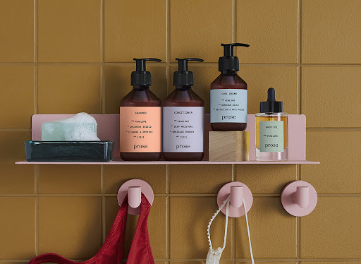 Shampoo, conditioner, hair oil, and soap with foam on a pink shelf against mustard-colored square tiled bathroom wall.