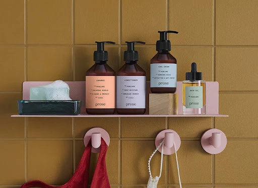 Shampoo, conditioner, hair oil, and soap with foam on a pink shelf against mustard-colored square tiled bathroom wall.