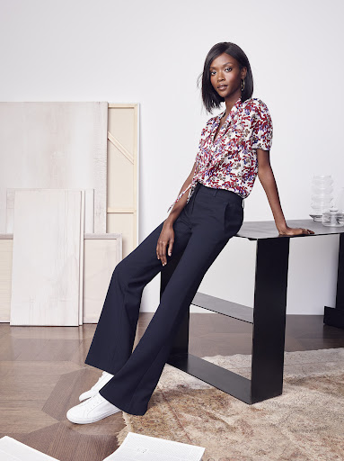 An adult woman poses in a floral blouse and flared trousers. She is sitting on a table in an indoor setting with studio lighting.