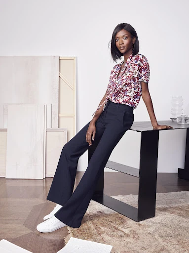 An adult woman poses in a floral blouse and flared trousers. She is sitting on a table in an indoor setting with studio lighting.
