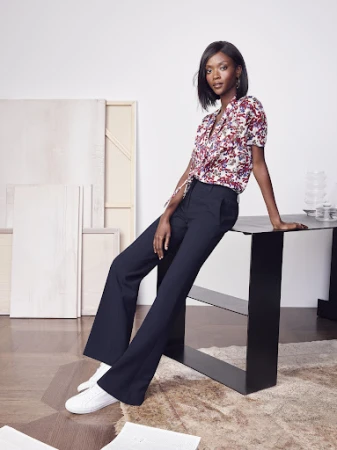 An adult woman poses in a floral blouse and flared trousers. She is sitting on a table in an indoor setting with studio lighting.