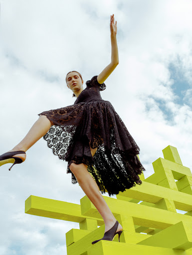 A young woman in a black lace dress strikes a dynamic pose atop a yellow geometric structure against a sky with scattered clouds.