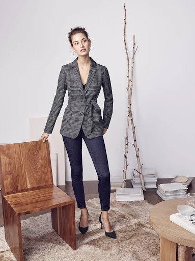 A woman poses in a gray plaid blazer, dark jeans, and heels indoors. A wood chair and decorative branches are in the background.