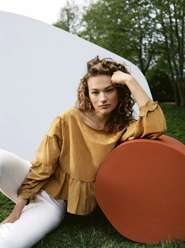 Portrait of a curly-haired woman in a yellow top and white pants posing against a white backdrop in a green grassy outdoor setting.