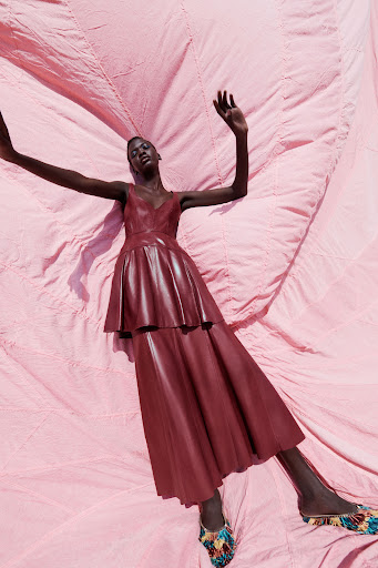 A Black female model wearing a burgundy dress lies on a pink fabric backdrop in an editorial fashion pose with arms raised.