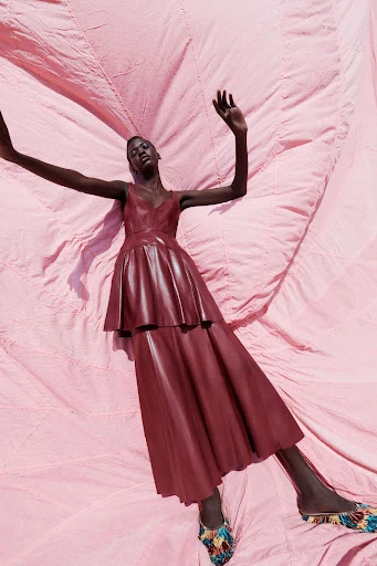 A Black female model wearing a burgundy dress lies on a pink fabric backdrop in an editorial fashion pose with arms raised.