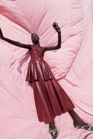 A Black female model wearing a burgundy dress lies on a pink fabric backdrop in an editorial fashion pose with arms raised.