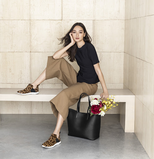 Woman in dark top and brown culottes sitting on bench with black tote bag and flowers, wearing leopard print sneakers.