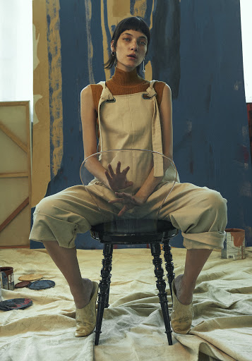 A young adult female in a casual jumpsuit sits on a stool in an art studio with paint cans and a backdrop covered in brushstrokes.