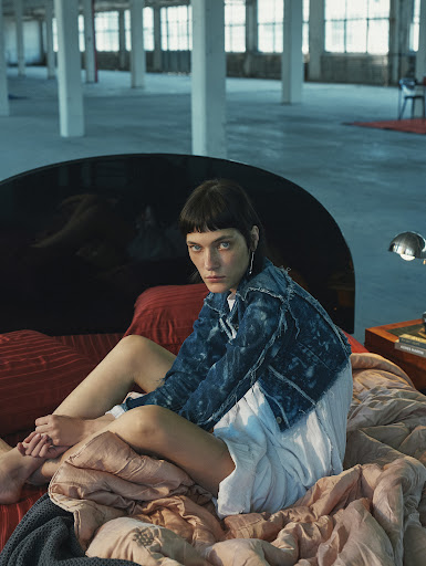 Young woman in casual clothing sitting on a bed in a large, industrial-style room with neutral tones and soft lighting, fashion editorial.