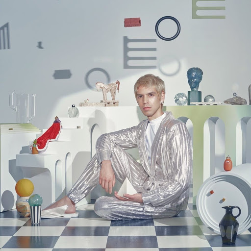 Young adult with blonde hair sitting cross-legged on a checkered floor in a studio setting with various objects in the background.
