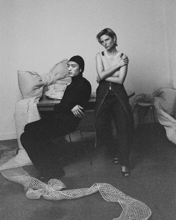Monochrome portrait of a man seated in a black turtleneck and a woman standing in dark trousers, styled by Lauren Bahr.