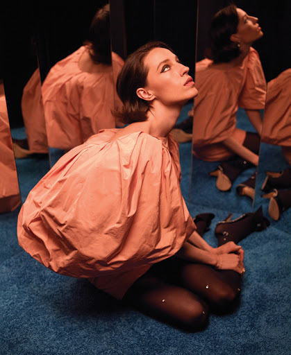 A stylish woman with dark hair kneels on a blue carpet, wearing a peach-colored dress, posing next to reflecting panels in a studio setting.