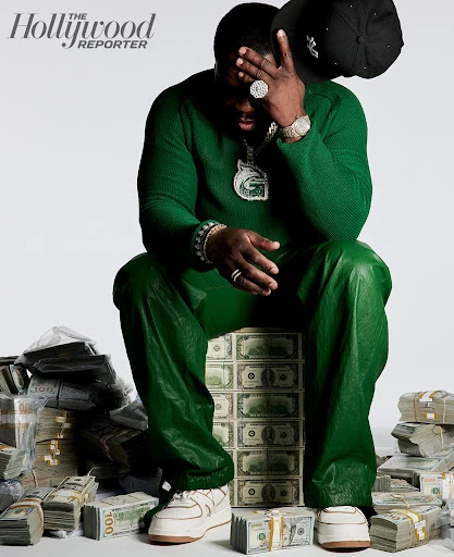 Man in green outfit poses seated on stacks of $100 bills, holding a baseball cap in photo studio with The Hollywood Reporter logo.