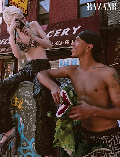 Two fashion models pose in front of a brick building; one holds a camera and the other holds a green stuffed dinosaur toy for a magazine shoot.