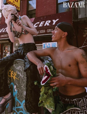 Two fashion models pose in front of a brick building; one holds a camera and the other holds a green stuffed dinosaur toy for a magazine shoot.