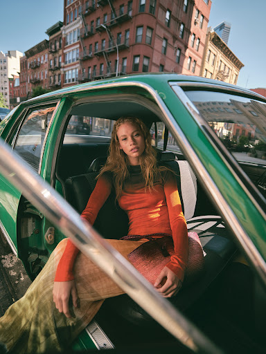 A woman with long curly hair wearing a gradient orange shirt and patterned pants is sitting inside a green car parked on a city street.