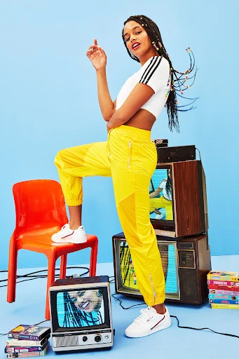Stylish woman poses with vintage televisions and VHS tapes in a vibrant studio setting, showcasing a blend of retro and modern fashion.