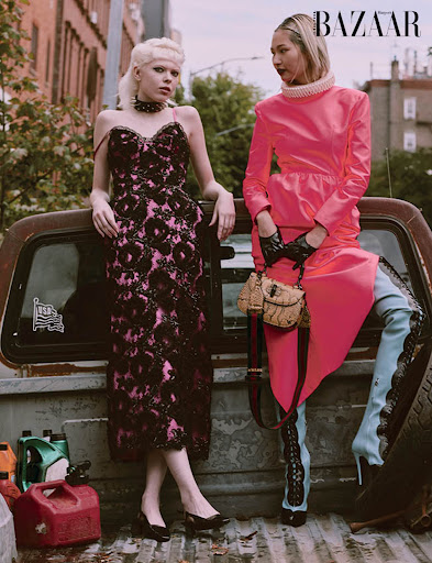 Two fashion models in dresses pose in a vintage pickup truck bed with gasoline cans, Harper's Bazaar logo visible, outdoor urban setting.