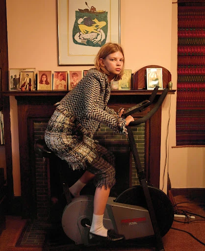 A young adult woman is riding an exercise bike in an indoor room setting. She is wearing a tweed suit and white socks with dark shoes.