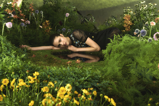 A young woman in a black dress with floral detail rests in a lush garden setting with vibrant yellow flowers and dark green foliage.